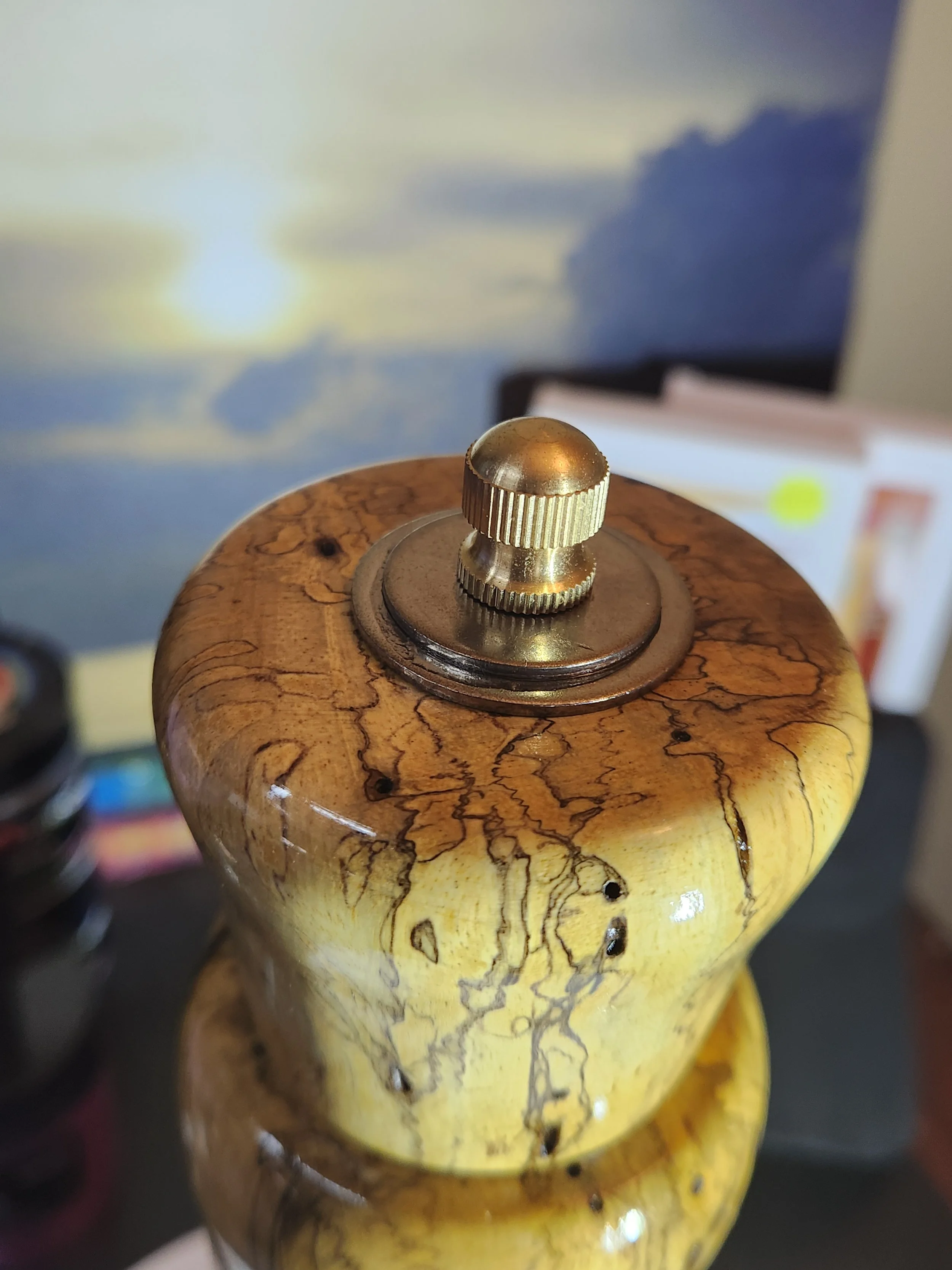 Close-up of a vintage wooden pepper mill with a brass knob on top, placed on a dark surface with a blurred background showing some papers and electronic devices.
