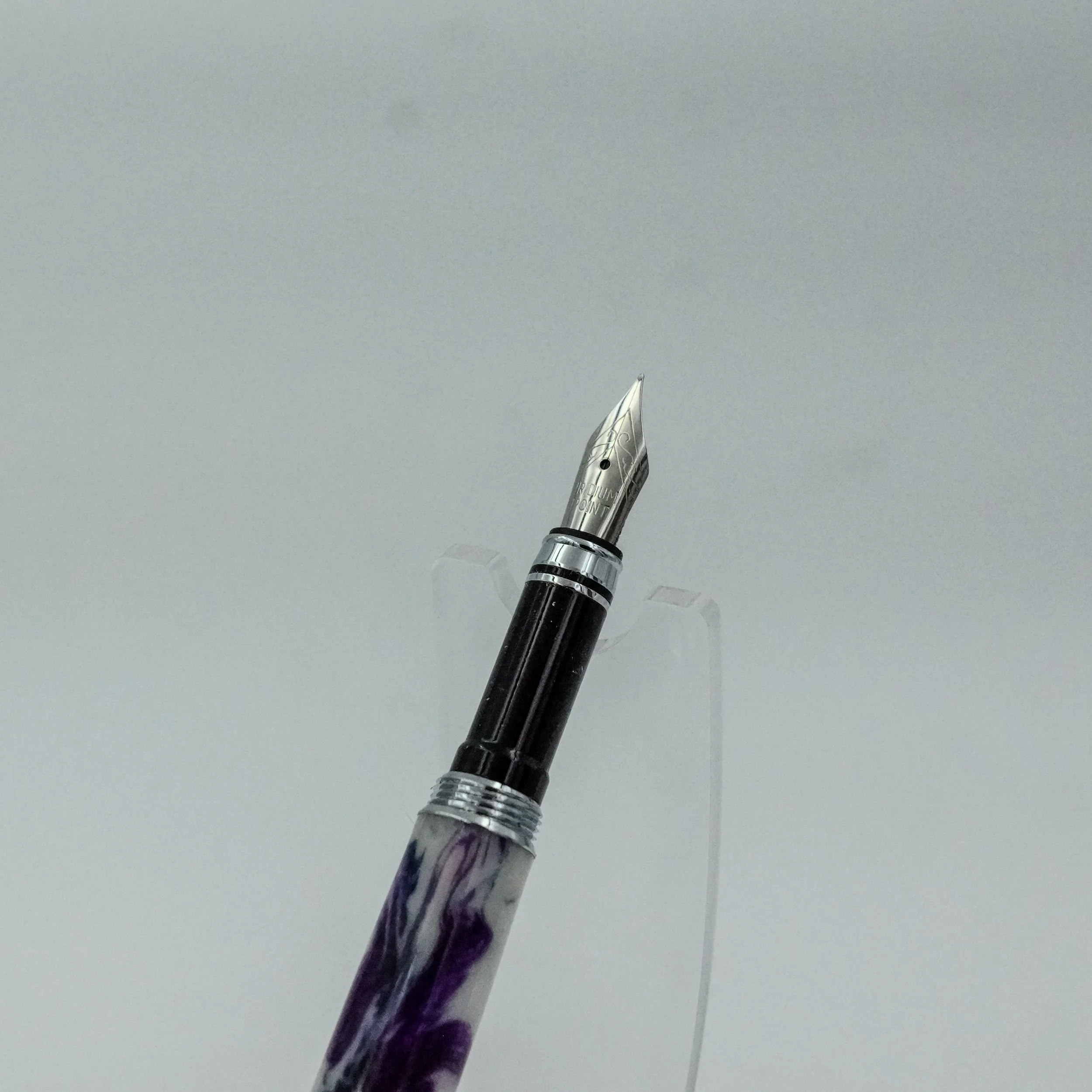 A close-up of a fountain pen with a marbled purple and white grip and silver accents, set against a plain light background.
