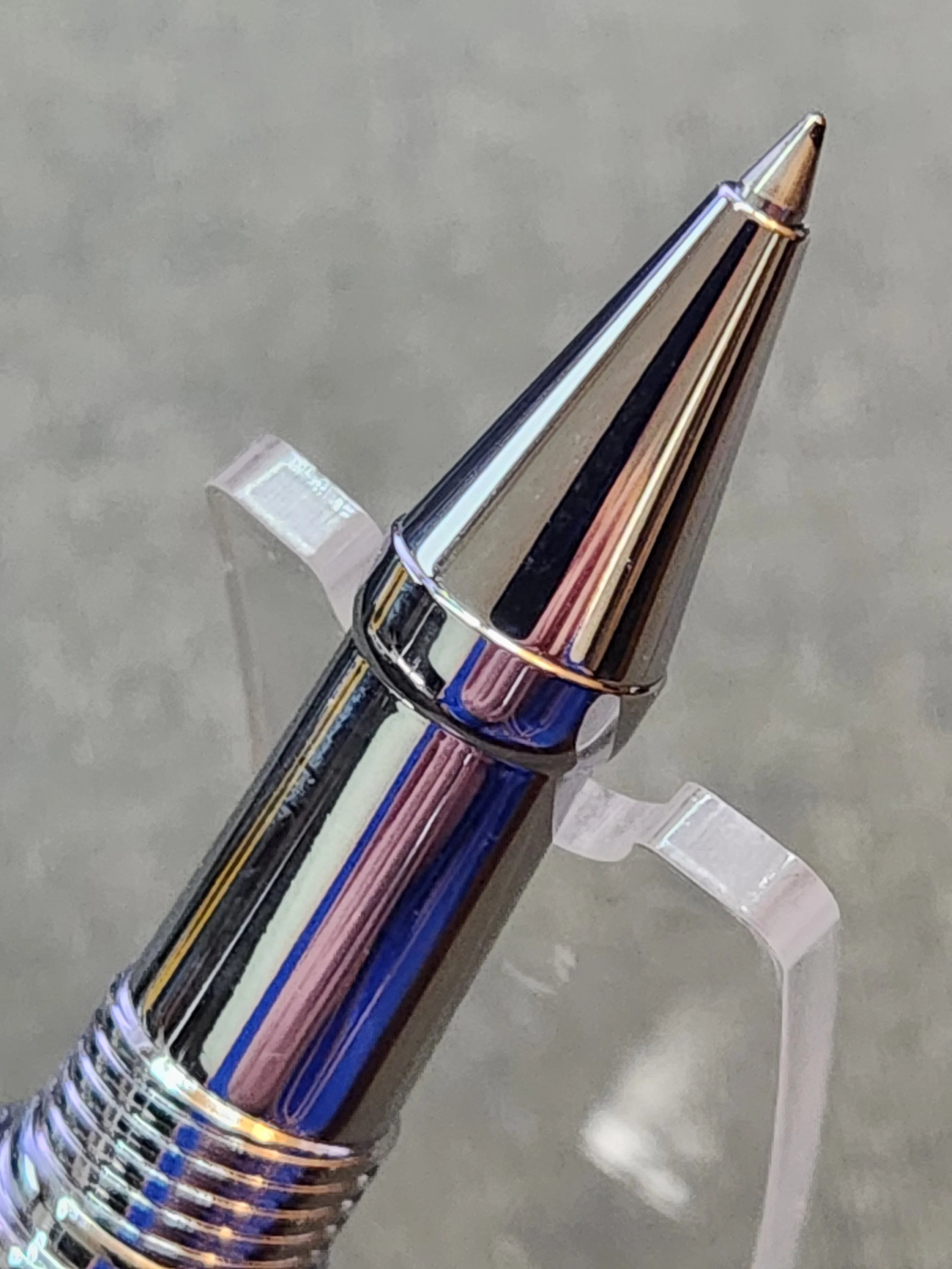 Close-up of a shiny metallic ballpoint pen with a gold tip, resting on a transparent stand against a gray background.