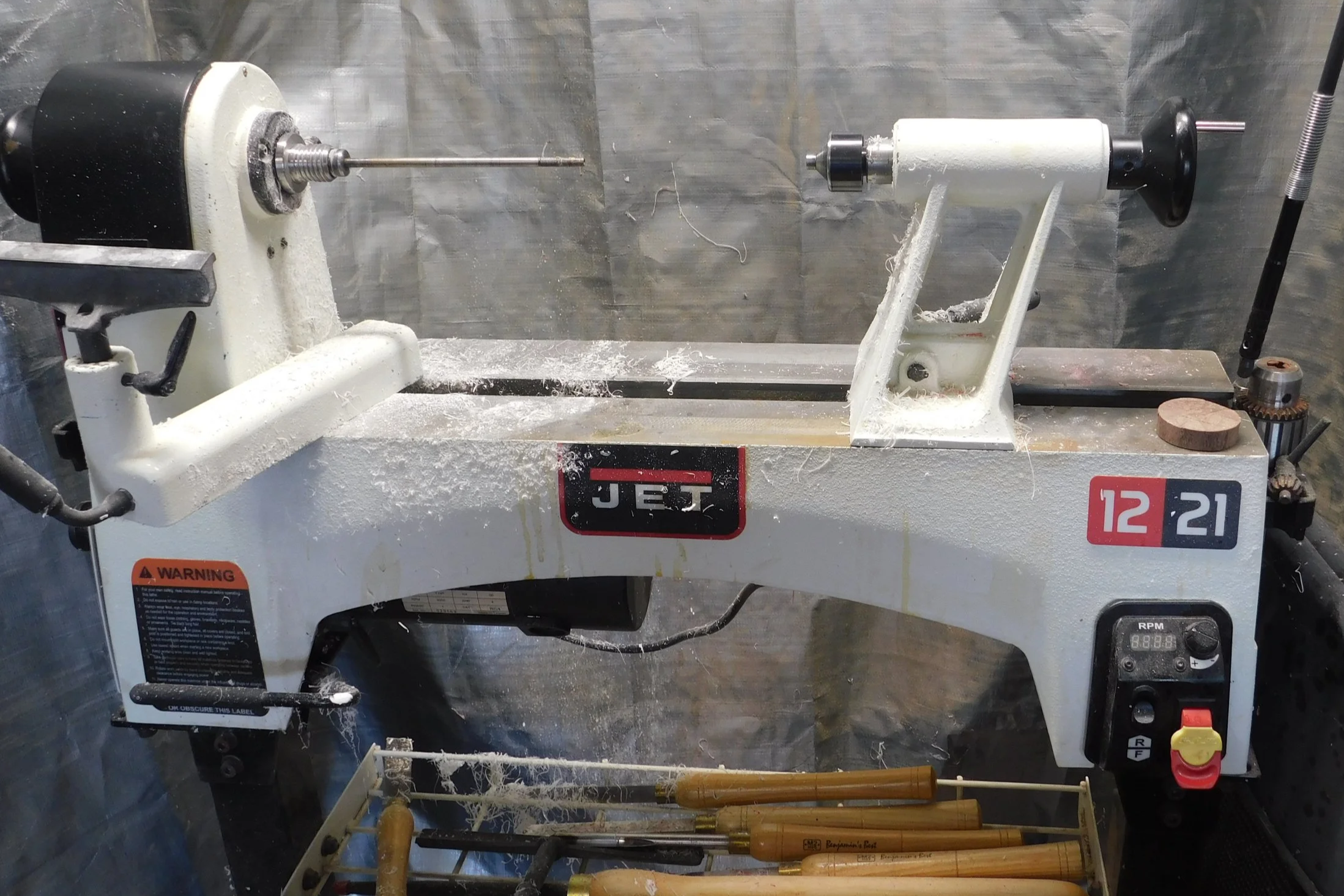 A small bench top lathe machine with the brand name JET and model 12-21, situated on a workbench, with several woodworking tools below it.