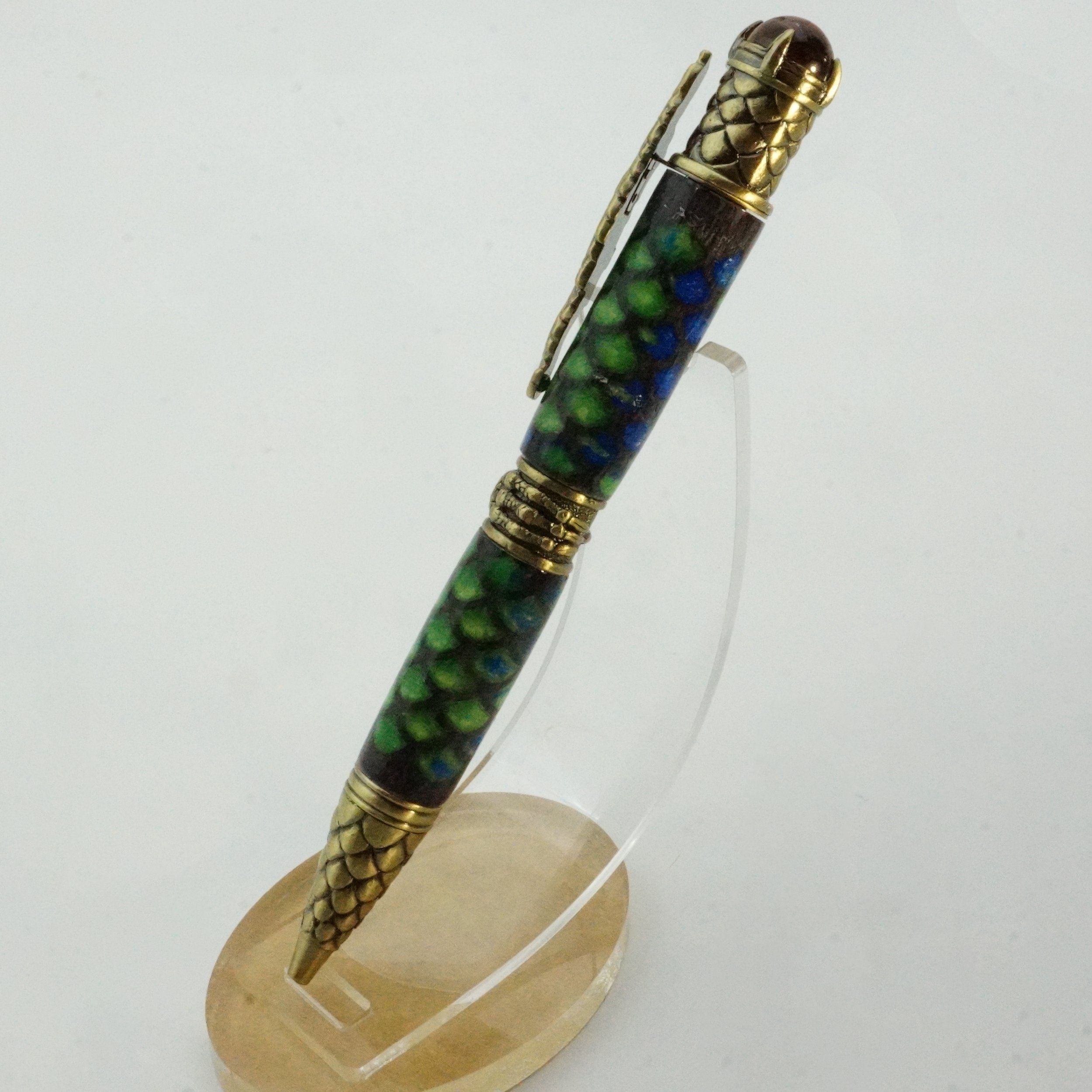 Decorative pen with gold-colored dragon scales at the top and bottom, and a multicolored marbled body, displayed on a clear stand.