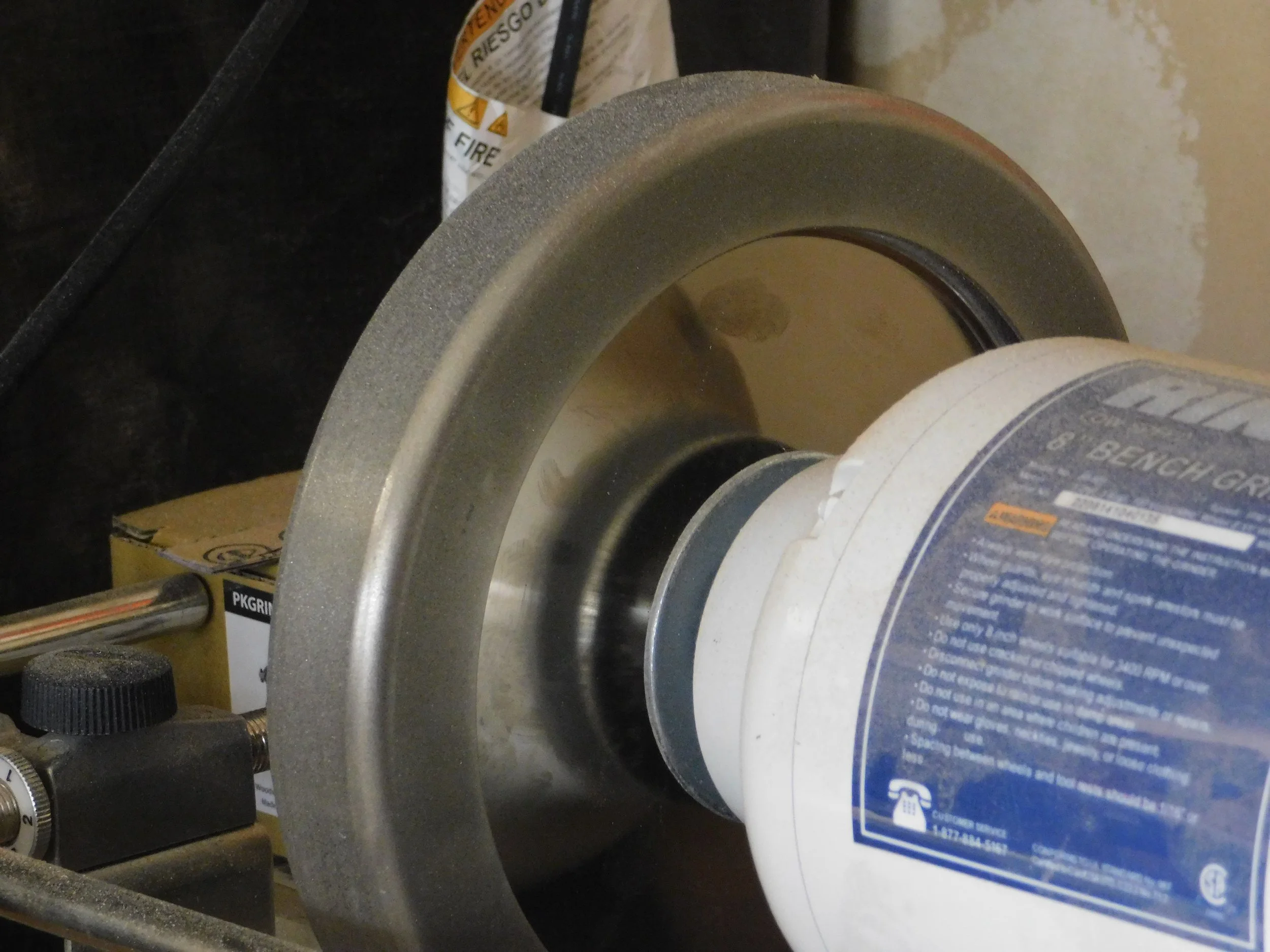 A metal grinder machine with a white plastic container on it. The container has blue and orange warning labels. The grinder has a large circular wheel and a handle, with other equipment and a cardboard box nearby.