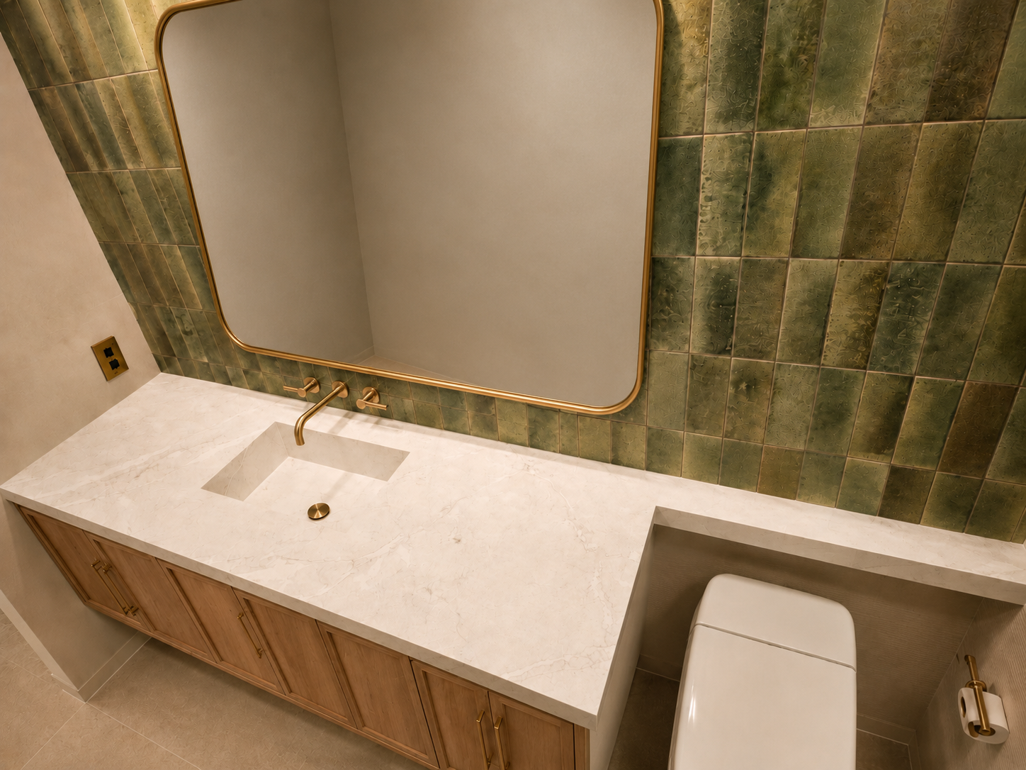 Bathroom sink with white marble countertop, gold fixtures, large mirror with gold frame, green tiled wall, and toilet.