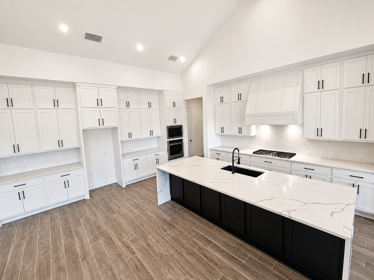 Modern kitchen with white cabinets, black hardware, a white marble island with black base, wood-look tile flooring, built-in oven and microwave, stovetop under a large vent hood, and a black sink with a black faucet.