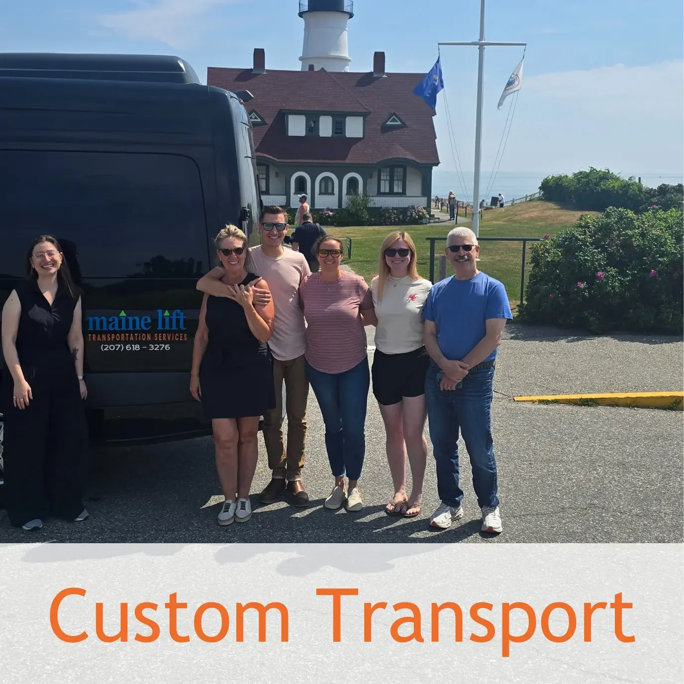 A group of six people standing outside near a black van with the logo 'Maine Lift Transportation Services' in front of a lighthouse and a house, with a cloudy sky.