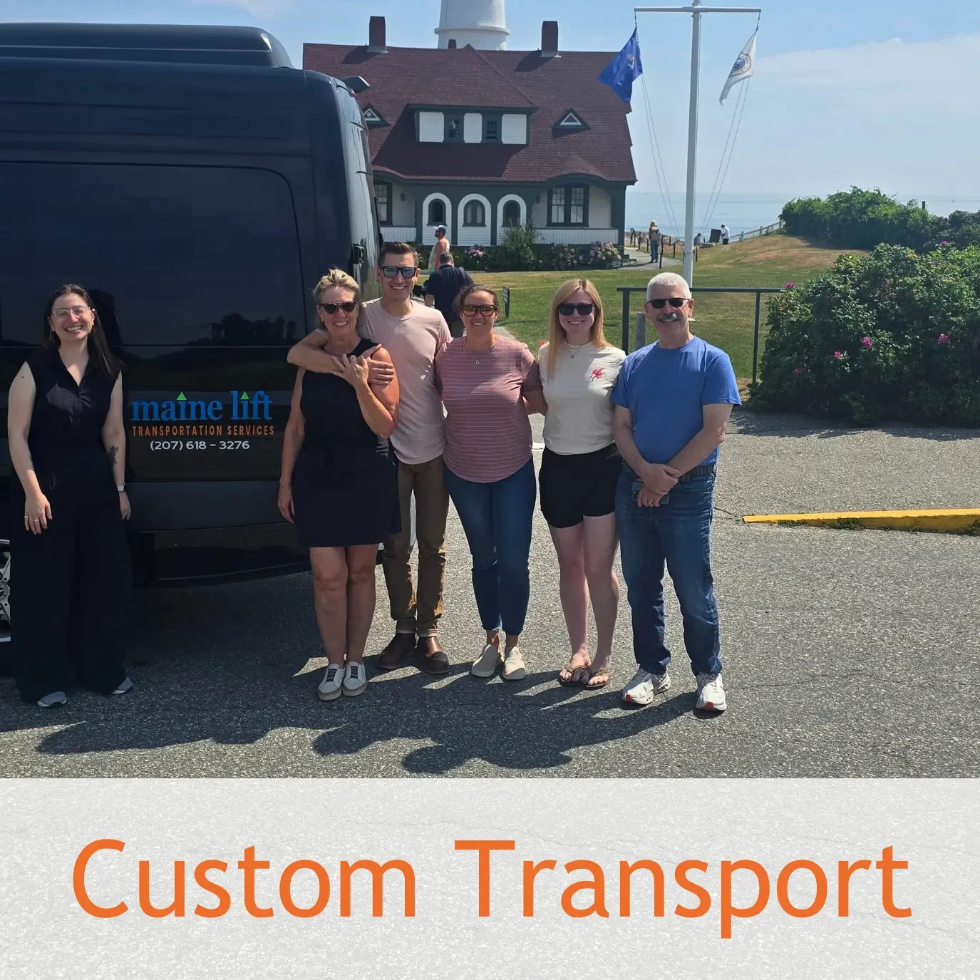 A group of six people standing in front of a black van with the text 'maine lift TRANSPORTATION SERVICES' and a phone number. They are outdoors near a house with flags, bushes, and water in the background, on a sunny day. The text 'Custom Transport' is at the bottom of the image.
