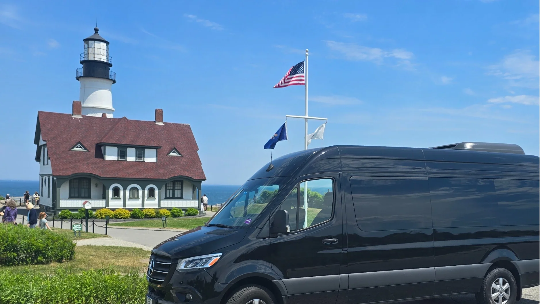 Lighthouse with a red roofed building nearby, American flags flying, and a black van in the foreground, against a clear blue sky and ocean background.