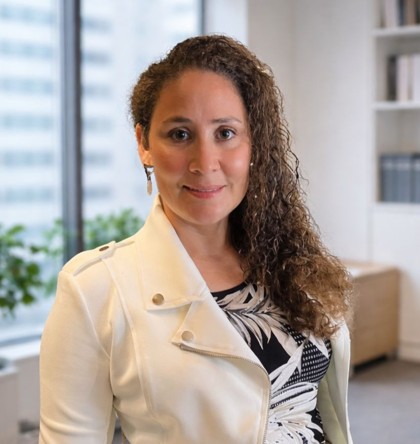 A woman with curly brown hair and earrings, smiling, indoors with a window showing city buildings behind her, wearing a white jacket and patterned top.