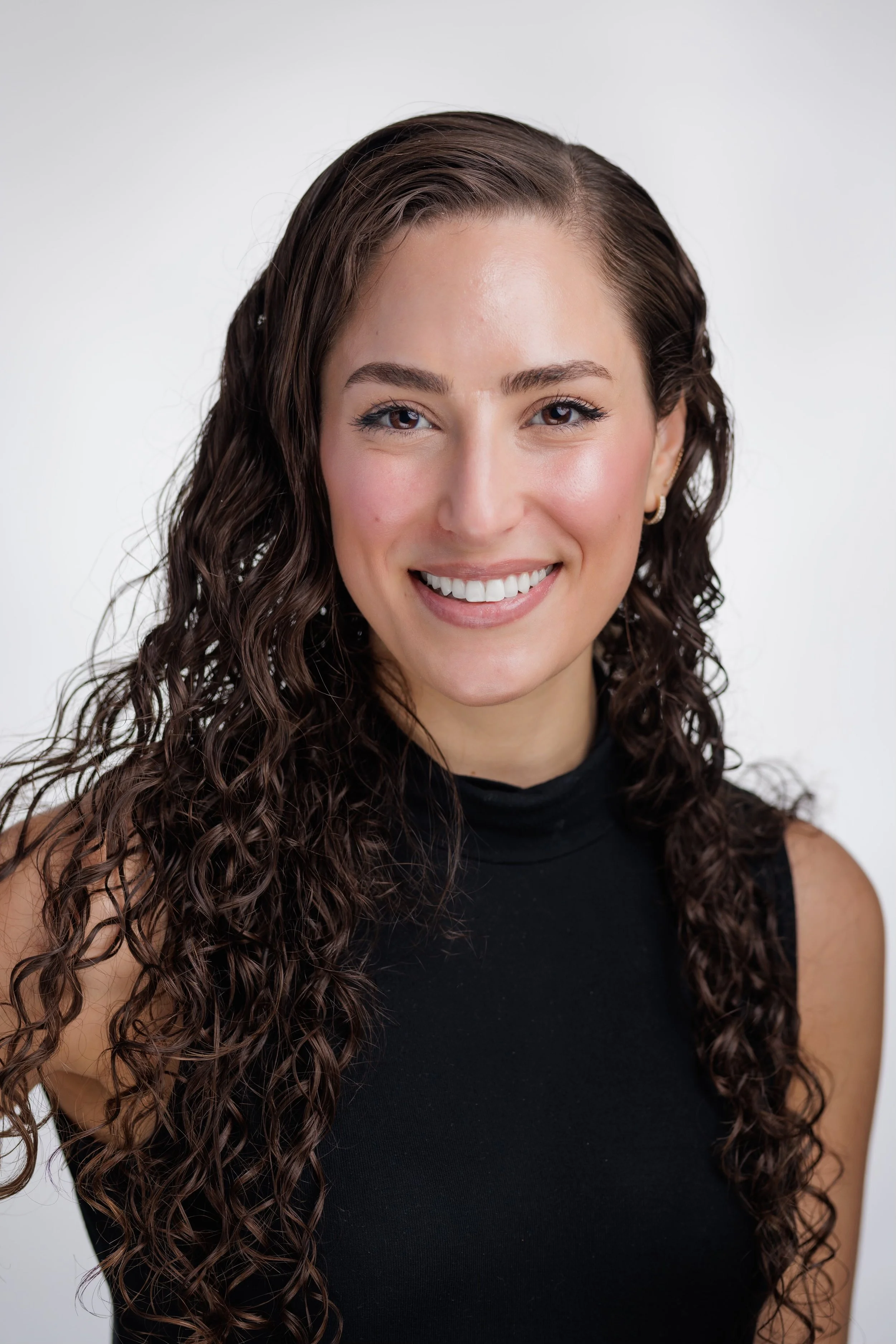 A woman with long, curly brown hair smiling, wearing a sleeveless black top, against a plain white background.