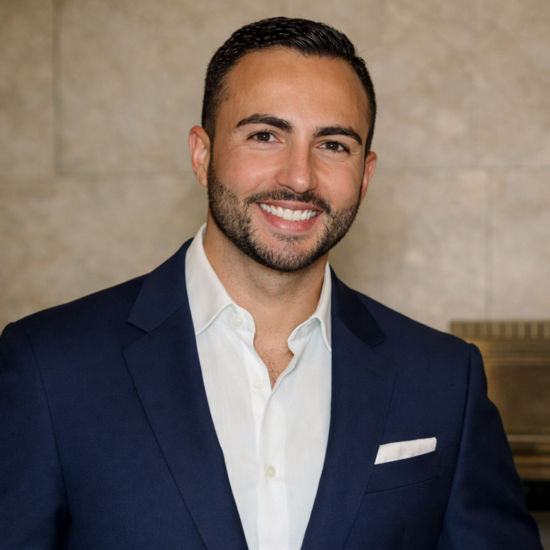 Professional portrait of a smiling man in a navy suit and white shirt, standing indoors against a beige wall.