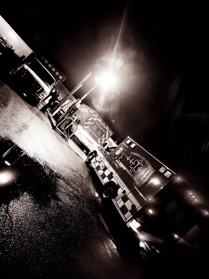 Nighttime scene of several emergency vehicles, including ambulances, parked on a wet road illuminated by streetlights.