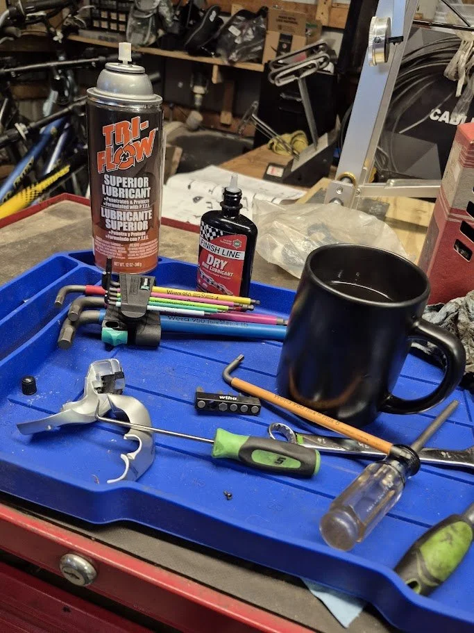 Tools and supplies scattered on a blue tray in a workshop, including a black coffee mug, a can of TRU-FLOW lubricant spray, a bottle of Finish Line dry lubricant, colorful zip ties, screwdrivers, an Allen wrench, and a bike chain link tool.