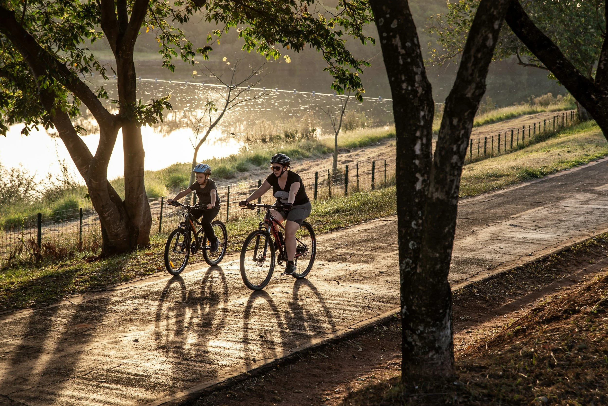 A woman and a child riding bicycles on a paved trail in a park by a lake with trees casting shadows on the ground during sunset.