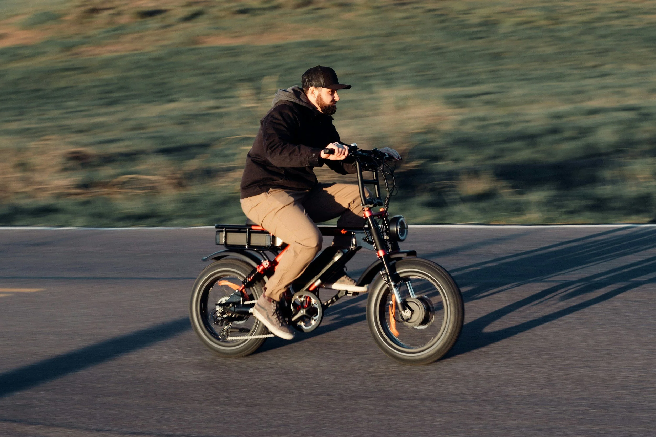 A man riding an electric bicycle on a road with grassy hills in the background during sunset.