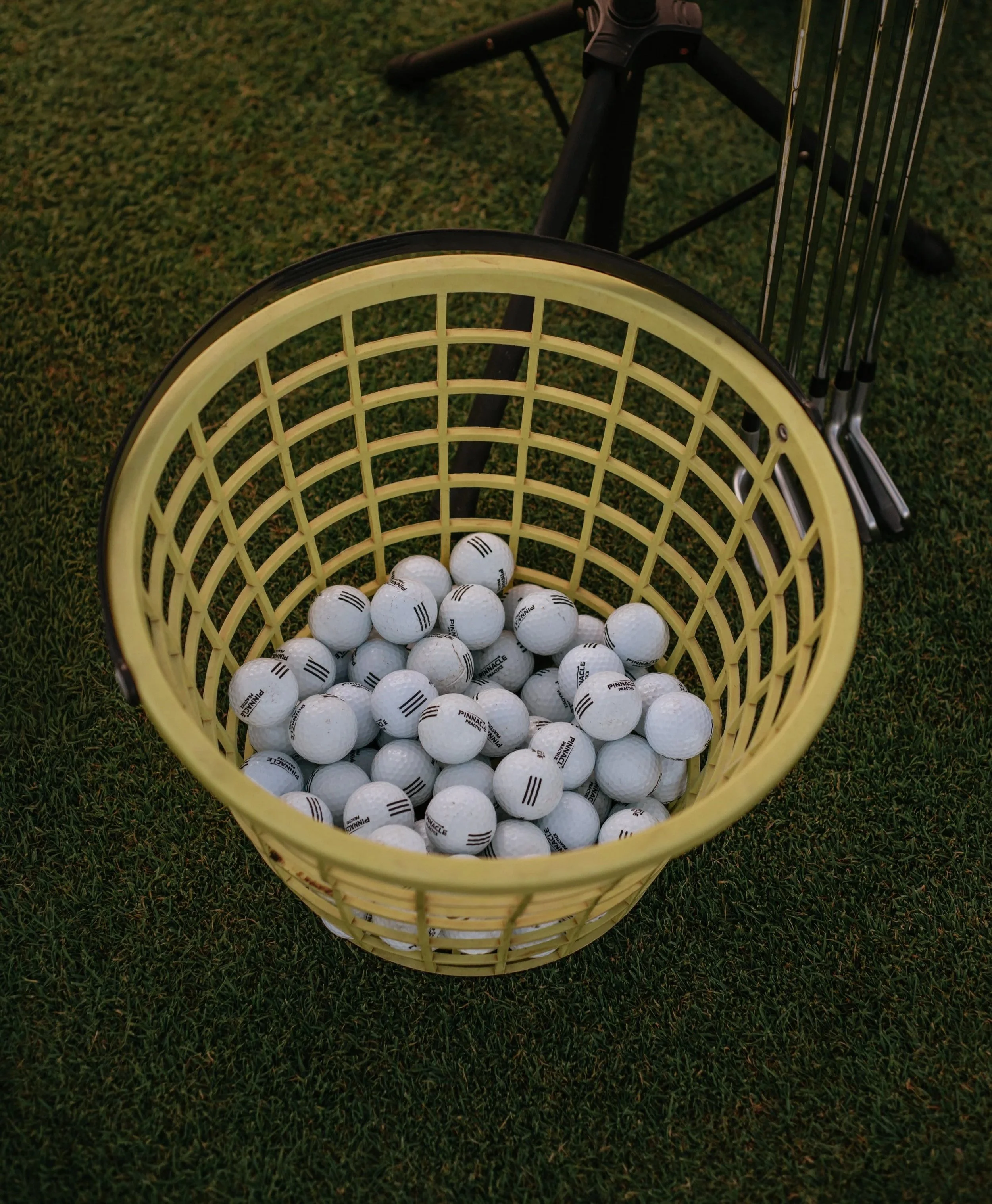 A yellow basket filled with white golf balls on grass with a golf club in the background.