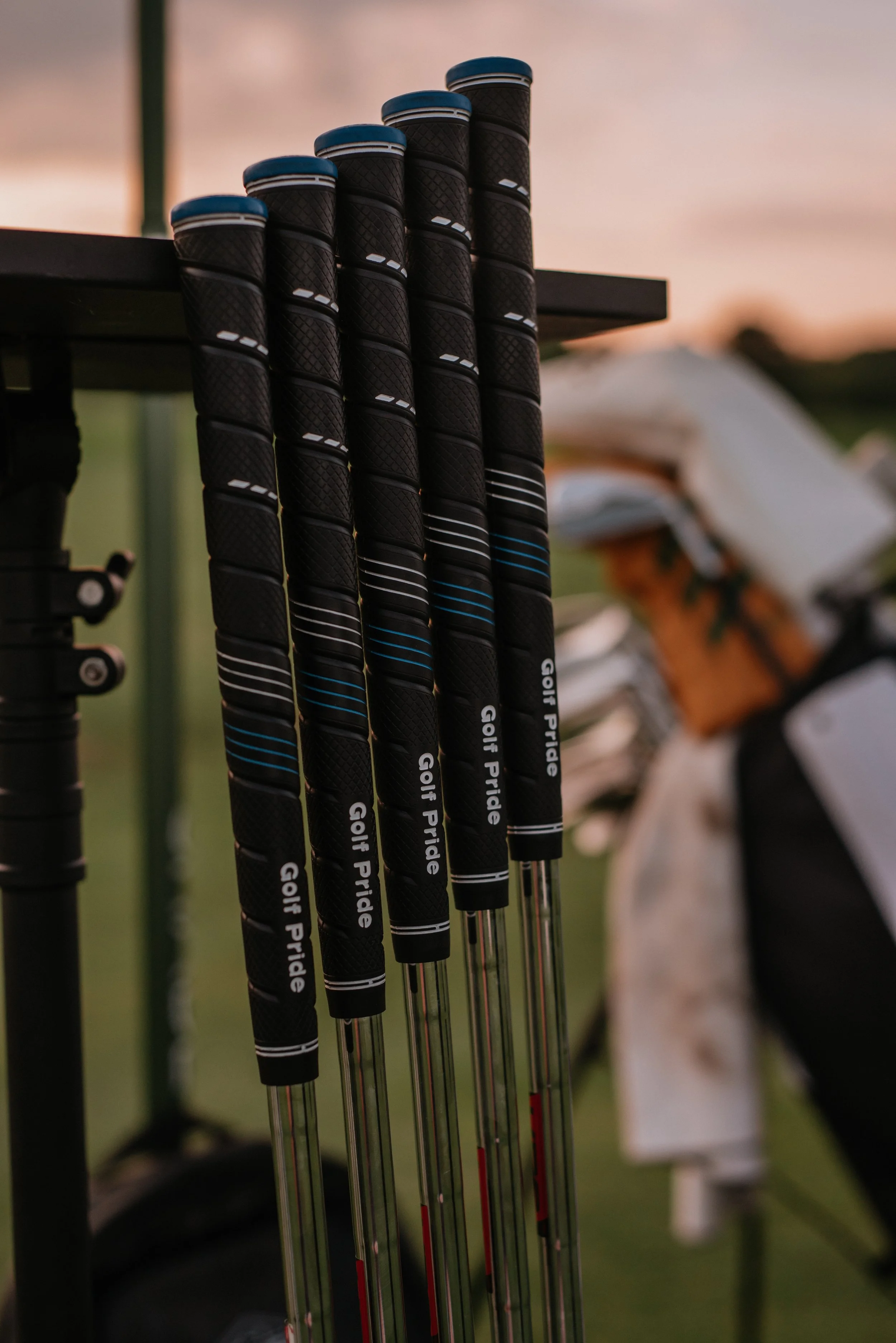 A row of six golf clubs with black grips and white and blue accents, standing upright in a golf bag or holder, during sunset or early evening with a blurred background.