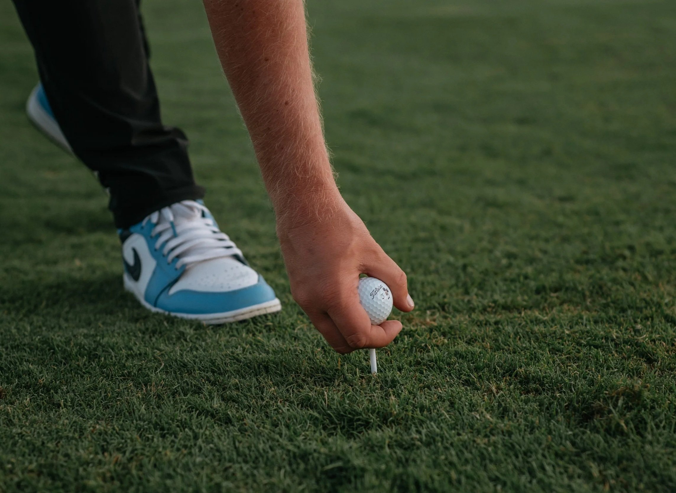 A person wearing black pants and white and blue sneakers is placing a golf ball on a tee on a golf course. The photo focuses on the person's arm, hand, and the golf ball.