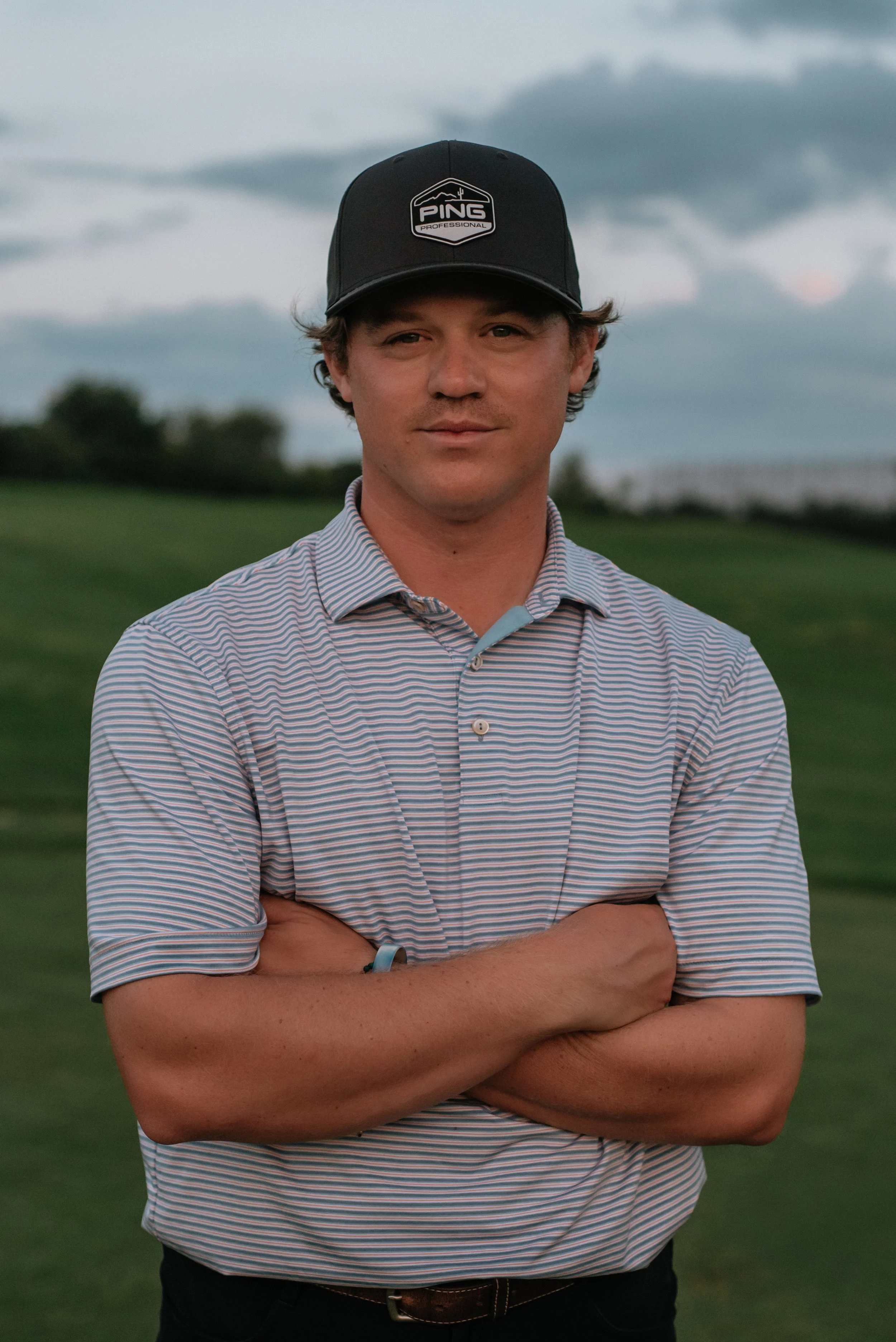 Patrick Cermak wearing a black PING golf cap and a striped polo shirt stands outdoors with arms crossed, looking at the camera. The background features a cloudy sky and a grassy field.