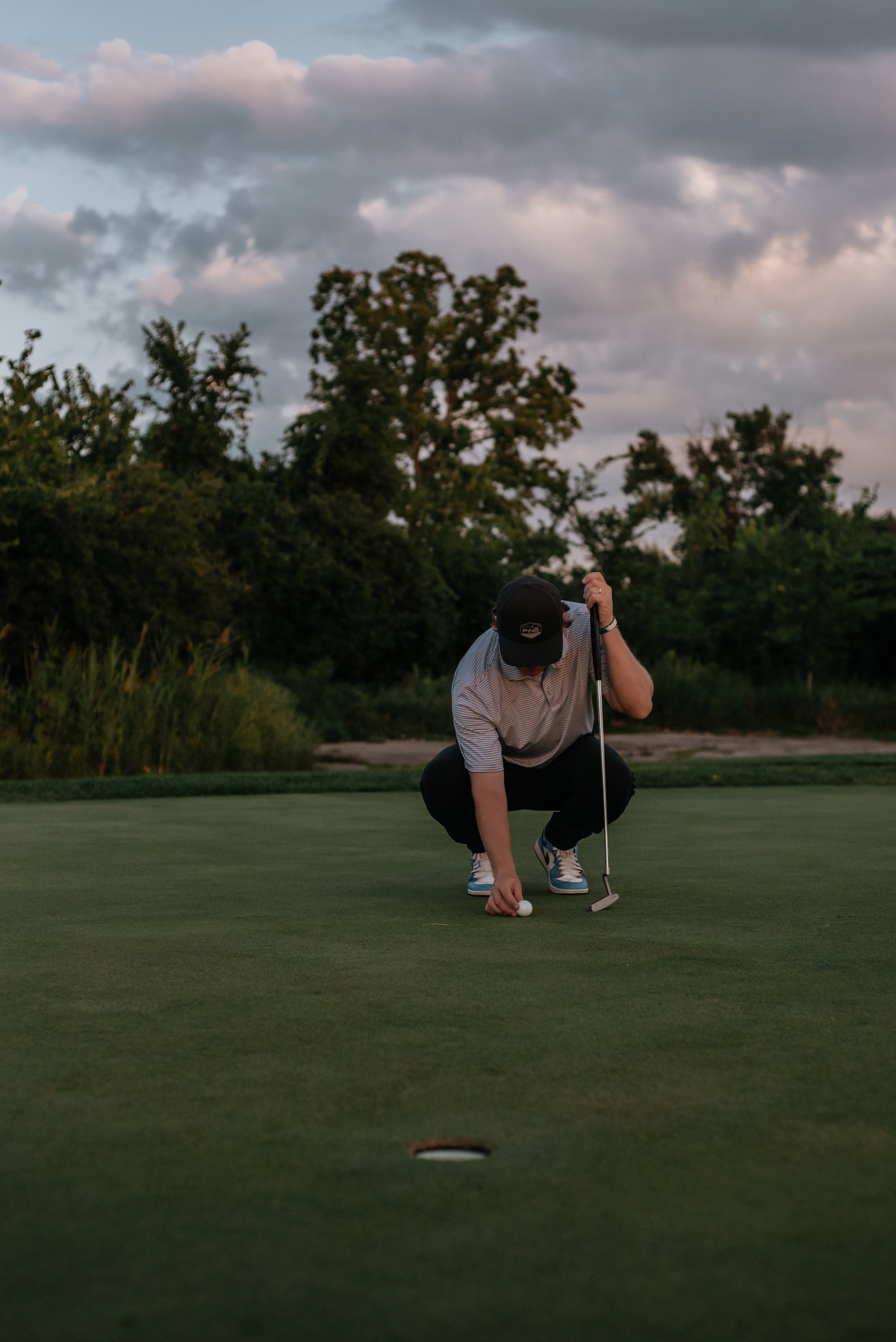 A person in a striped shirt, black cap, and sneakers is crouched down on a golf green, lining up a putt with a golf ball and putter during sunset, with trees and a cloudy sky in the background.