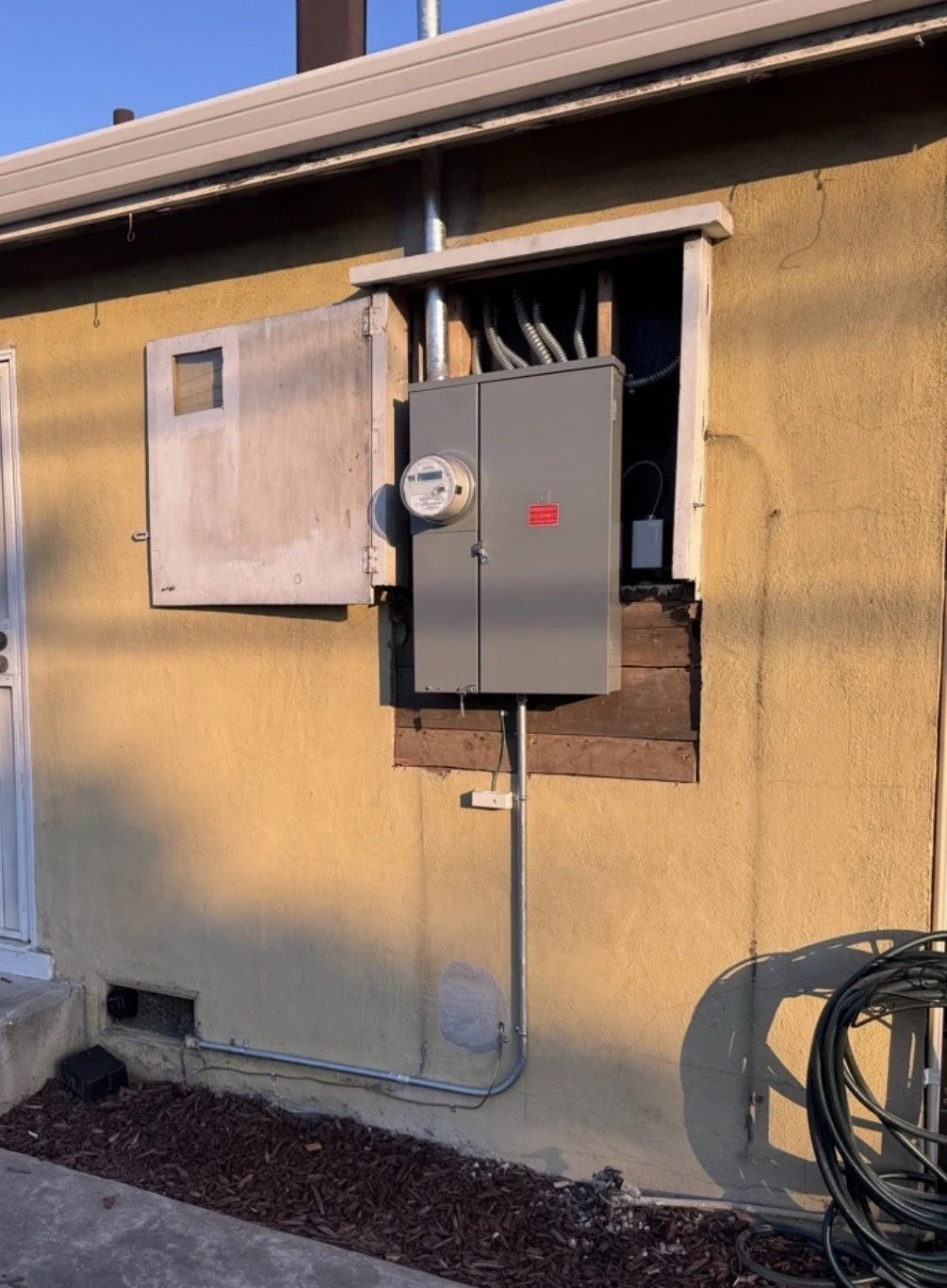 Exterior wall of a house with an electrical meter, a gray electrical panel, and conduit pipes.