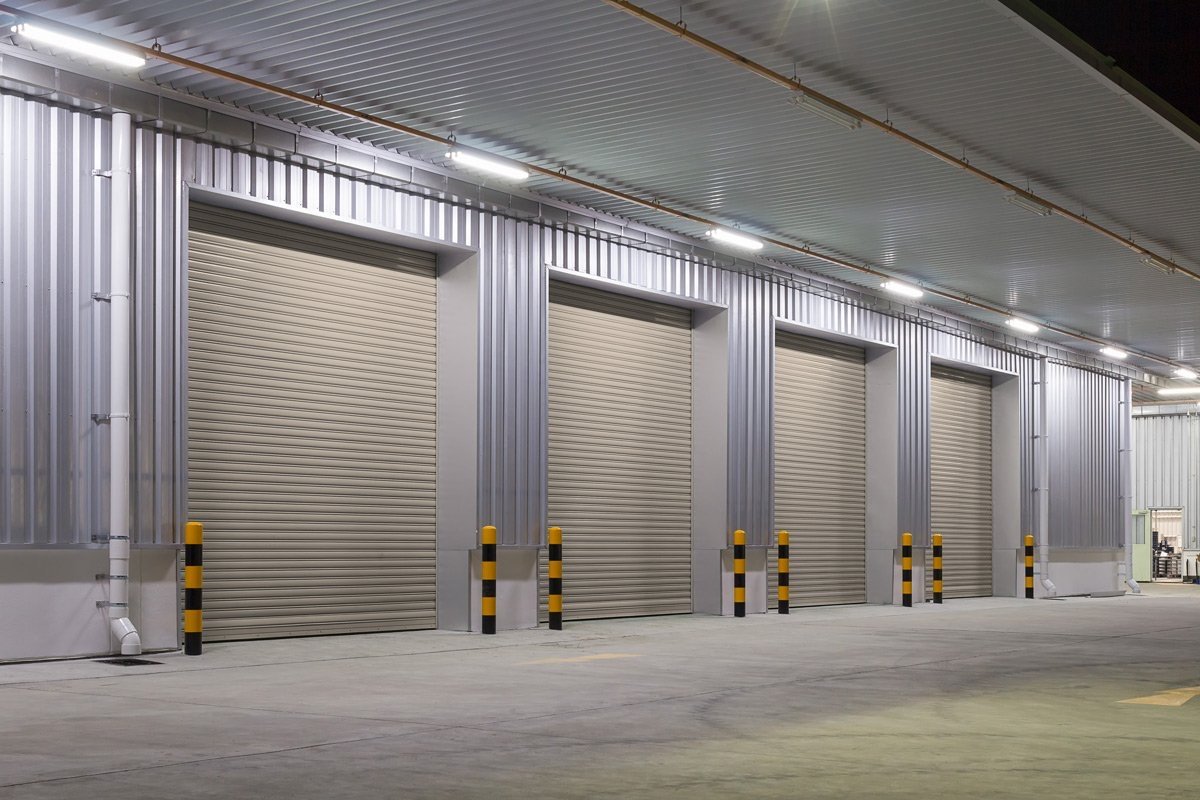 Empty industrial warehouse with four closed roll-up doors and yellow-black safety bollards in front.