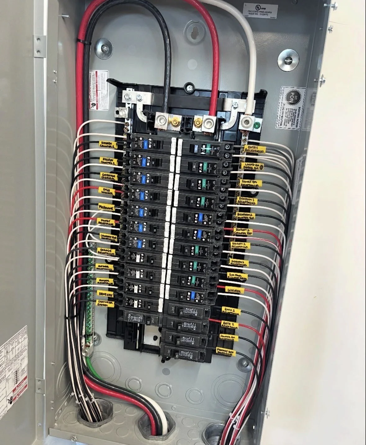 Electrical breaker panel with multiple circuit breakers, each labeled for different household circuits, with black, red, and white wiring connected.