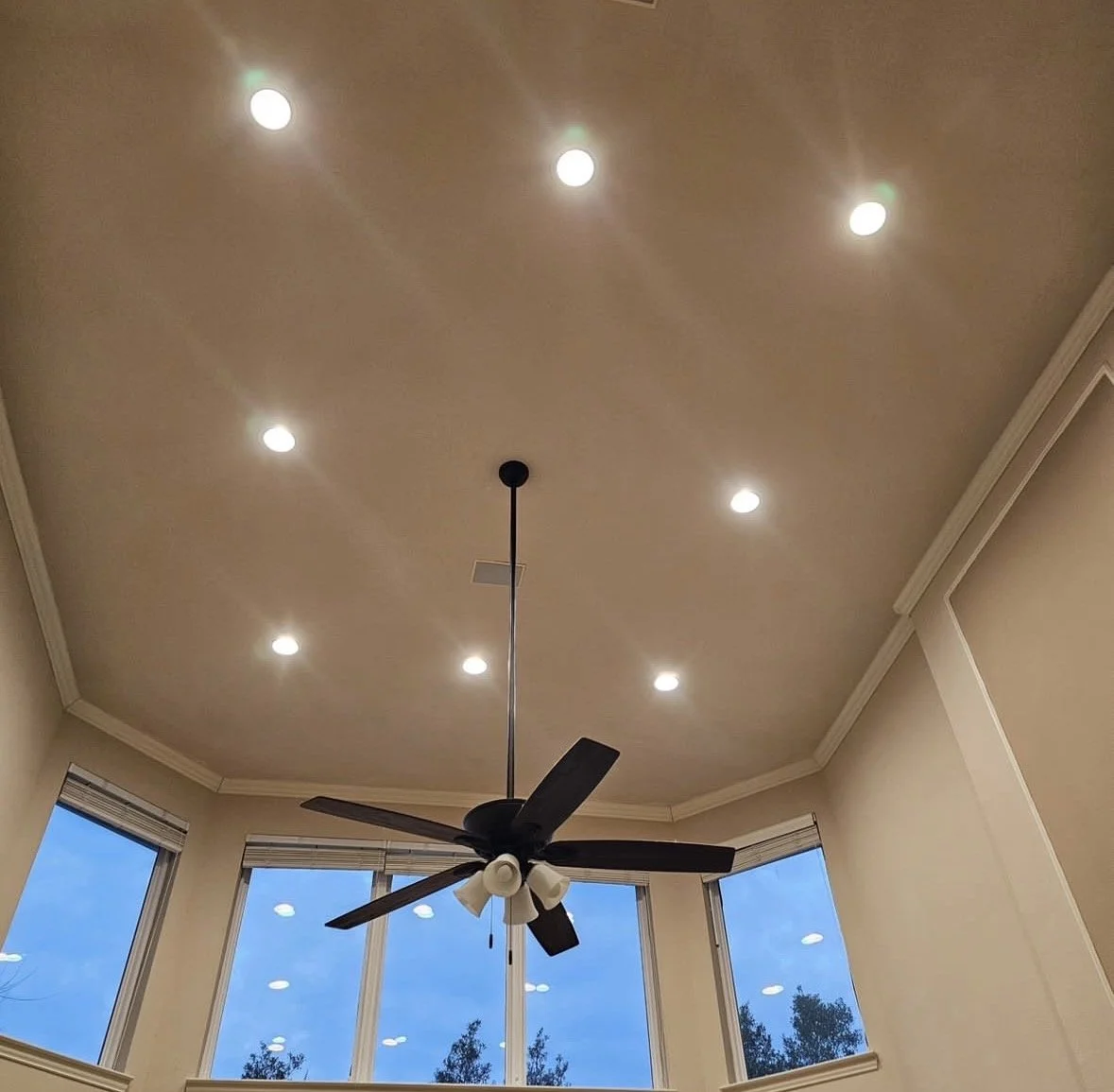 Ceiling with recessed lights, ceiling fan with five blades, and large windows showing a blue sky and trees outside.