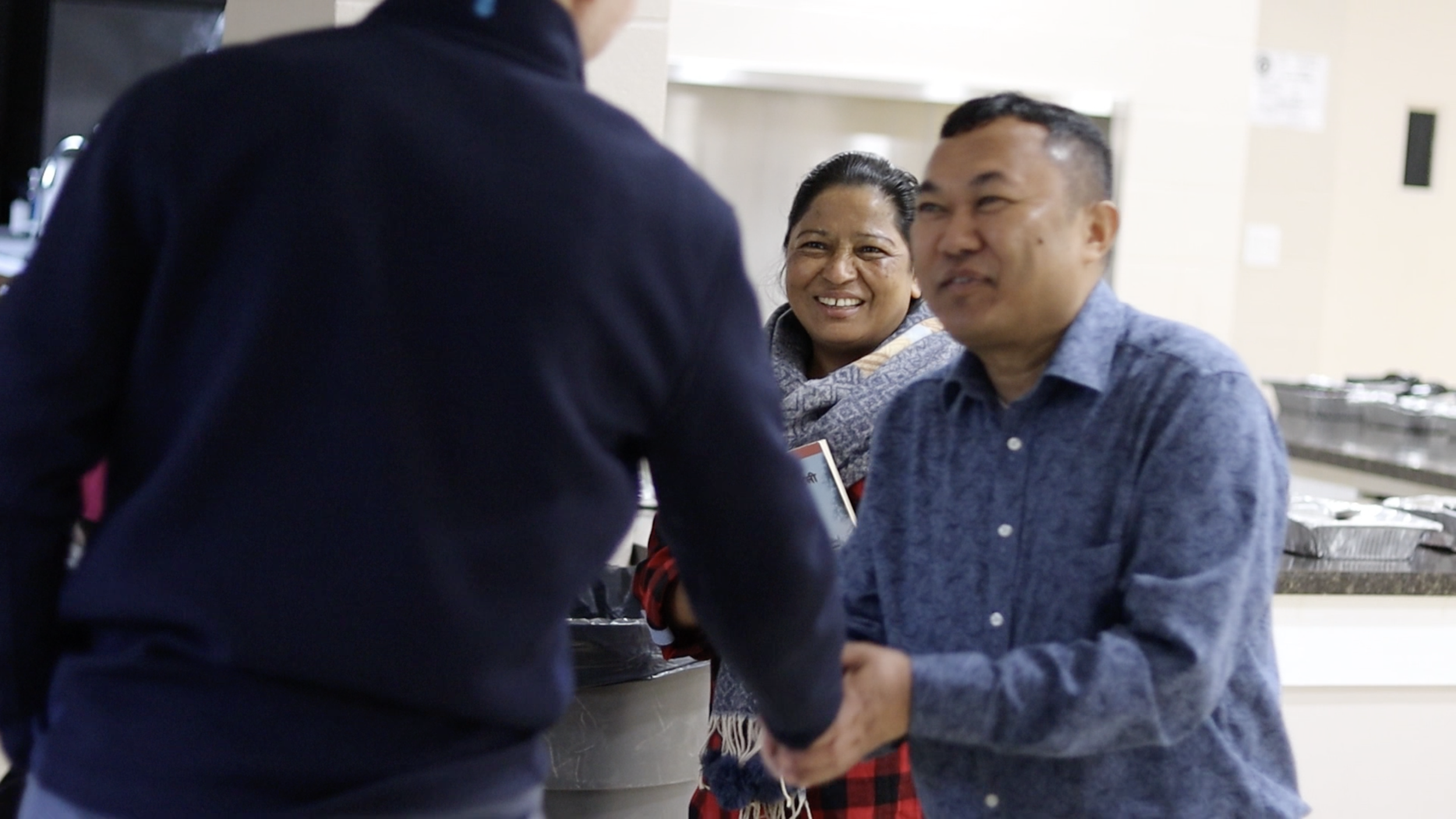 Two men and a woman smiling while interacting in a cafeteria or food service area.