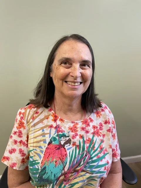 A woman with shoulder-length dark hair and a floral shirt with a parrot and tropical plant print, smiling at the camera against a plain light-colored wall.