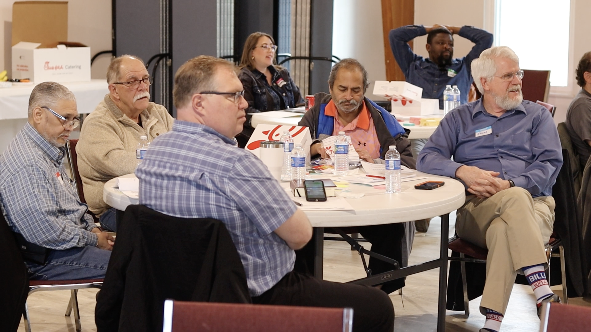 People attending a meeting or conference seated at round tables with papers, water bottles, and drinks; some are listening, and one person in the back has hands behind head.