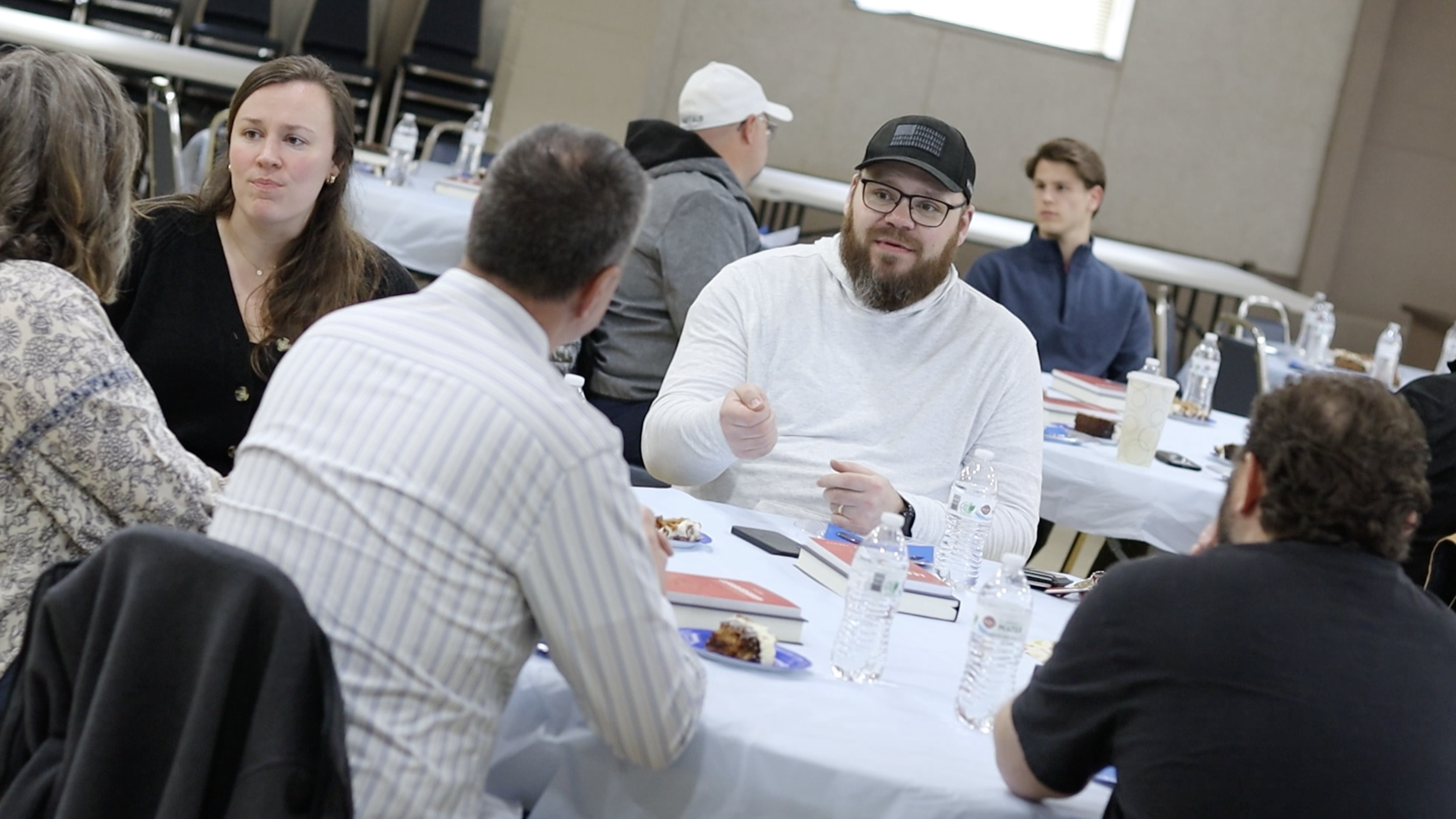 People sitting around a table engaged in conversation at a meeting or conference, with food and drinks present.