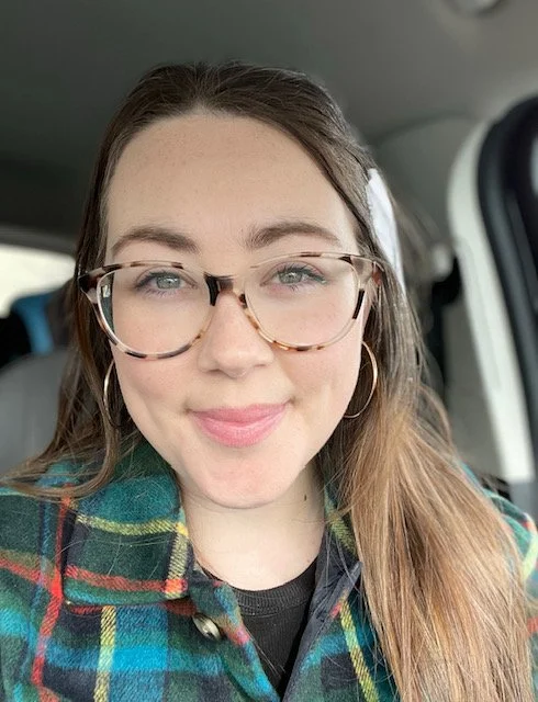 A young woman with glasses and long brown hair taking a selfie inside a vehicle, wearing a plaid shirt.
