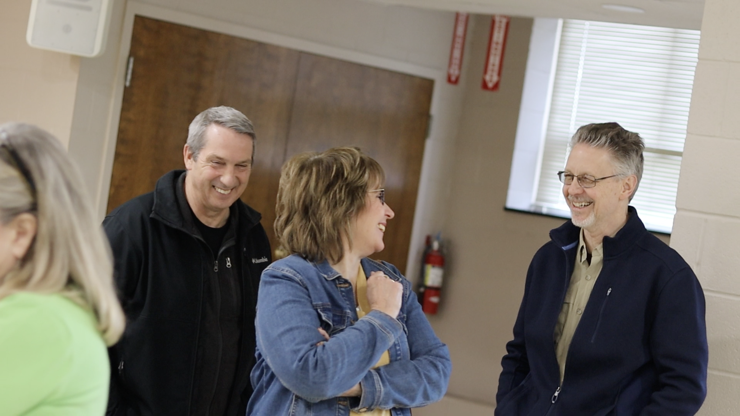 Group of four adults smiling and talking indoors, with two men on the left and right, and a woman in the middle, in a casual setting with window blinds and a fire extinguisher in the background.