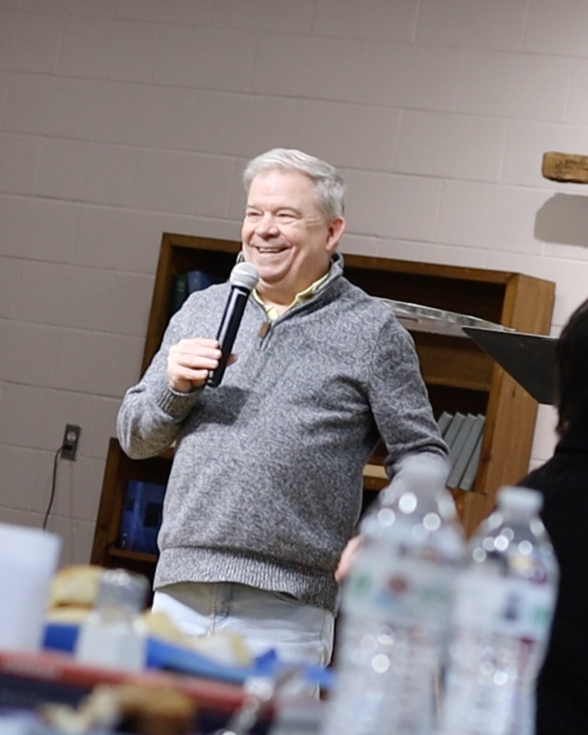 A man with gray hair smiling and holding a microphone, standing in a room with beige walls and a wooden bookshelf in the background.