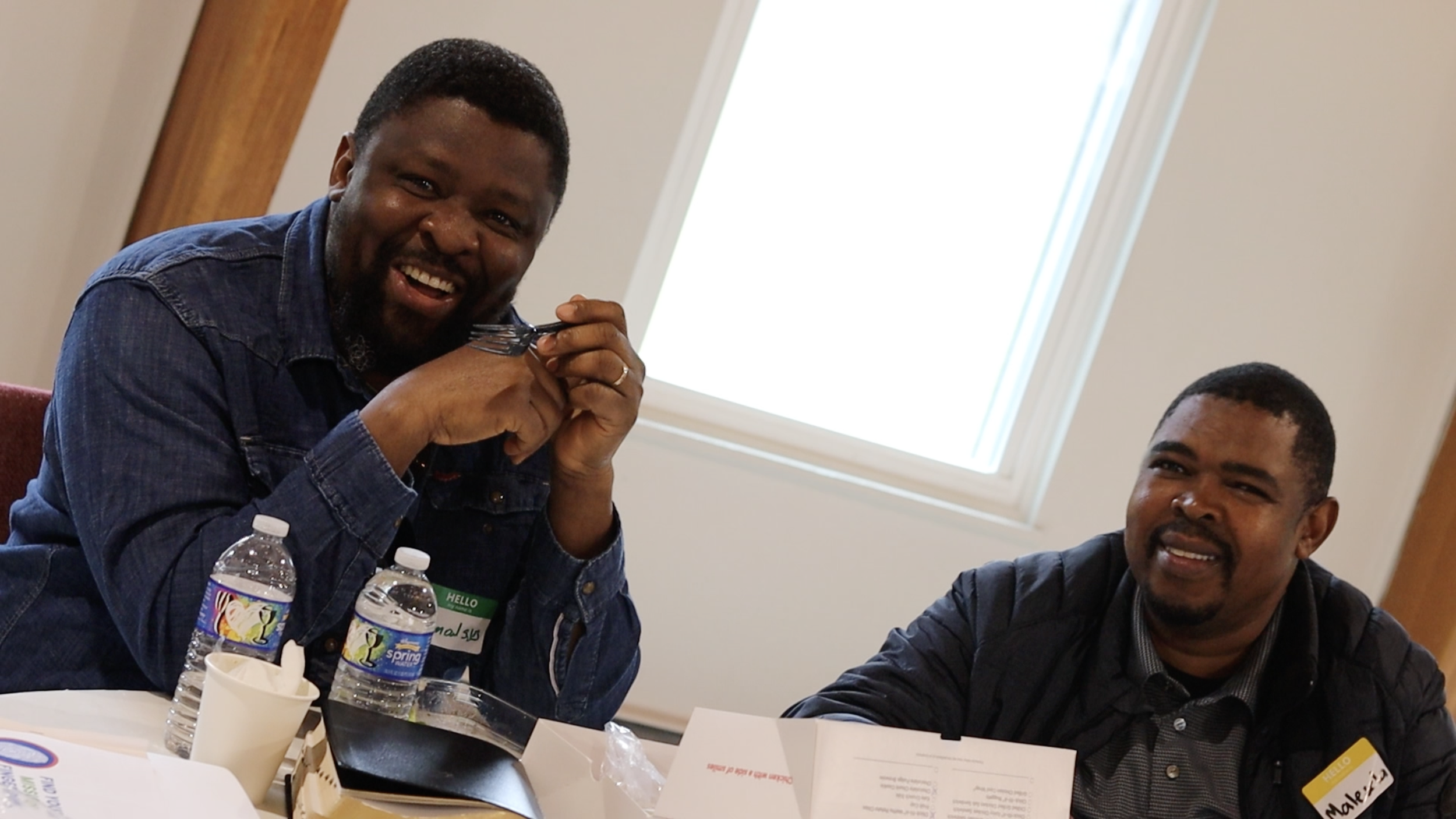 Two men sitting at a table, one smiling and holding a fork, with bottles of water and papers in front of them, in a brightly lit room with a skylight.