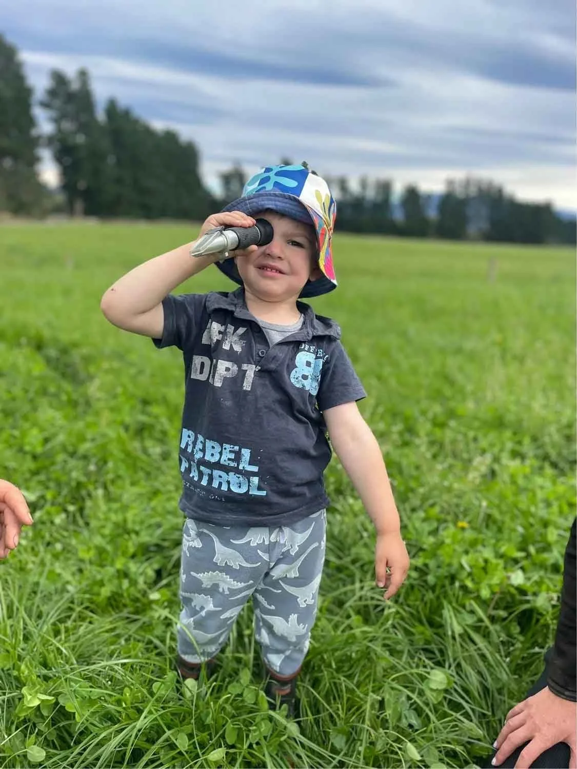 A young boy wearing a colorful hat and camouflage pants is holding a toy telescope to his eye in a grassy field. His expression appears curious or puzzled, and the background shows a cloudy sky and a distant tree line.