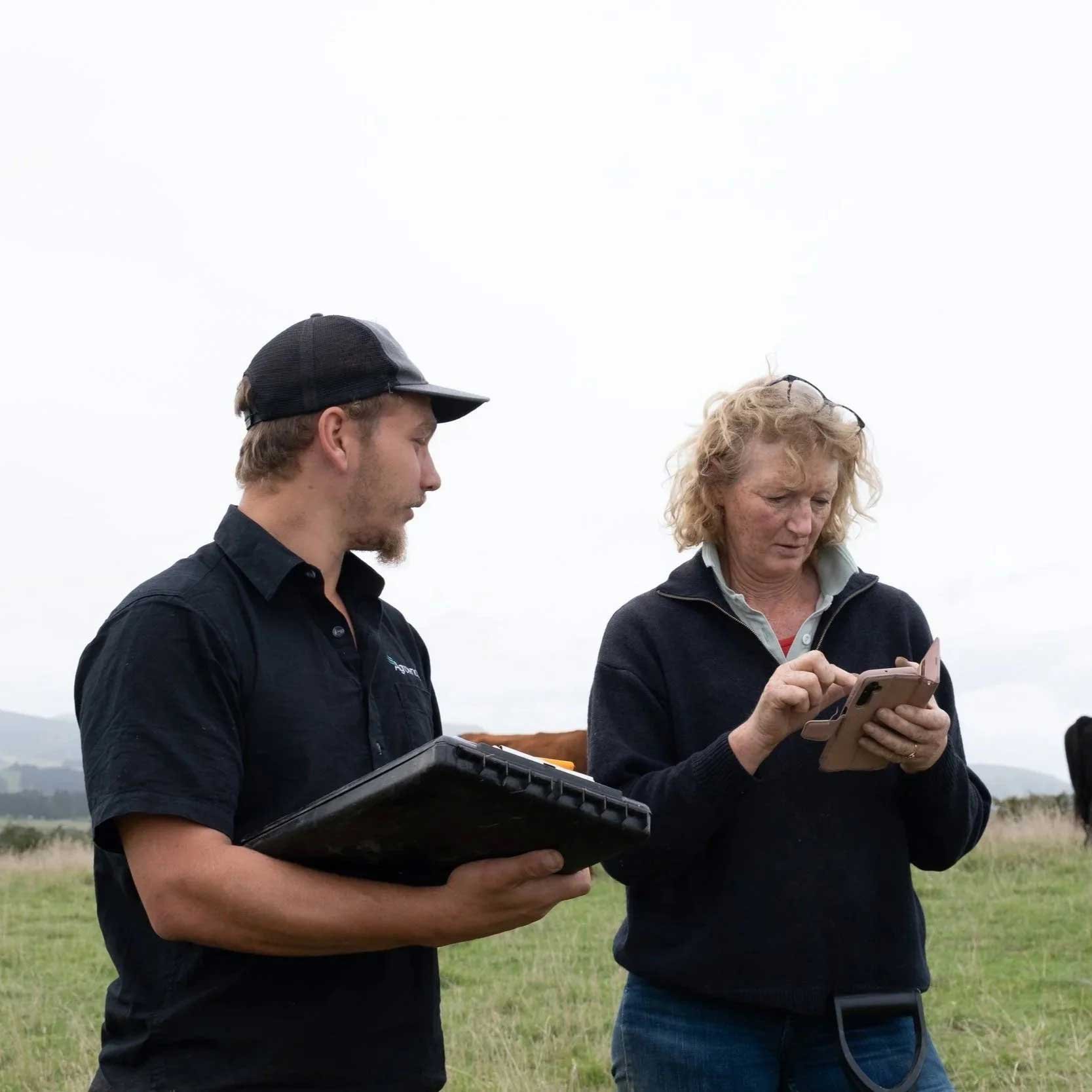 Two people, a man and a woman, standing outdoors in a farm paddock, looking at a smartphone and holding a tablet.