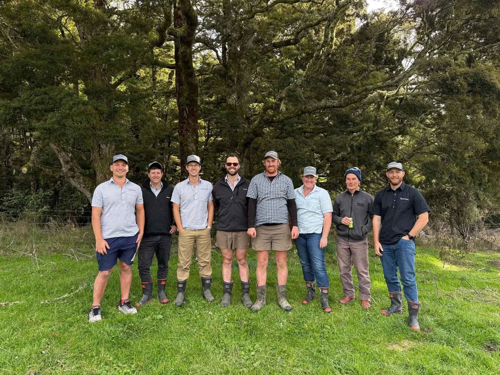 The Agrownomics team standing outdoors in front of a large tree, all smiling and dressed casually, some with hats and boots, on a grassy area.