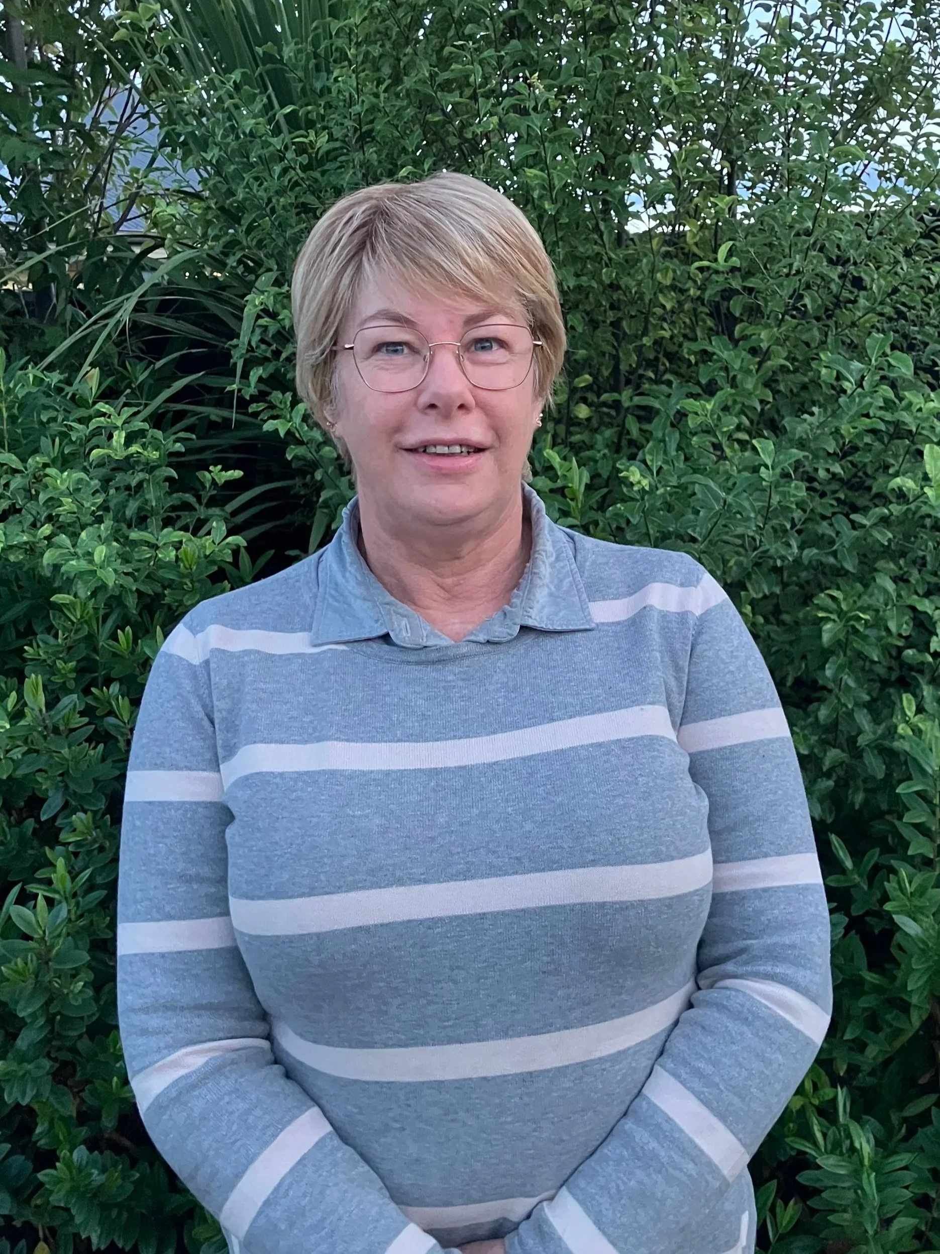Linda Sim Agrownomics Admin & Accounts, wearing glasses and a gray and white striped sweater standing outdoors in front of green bushes.