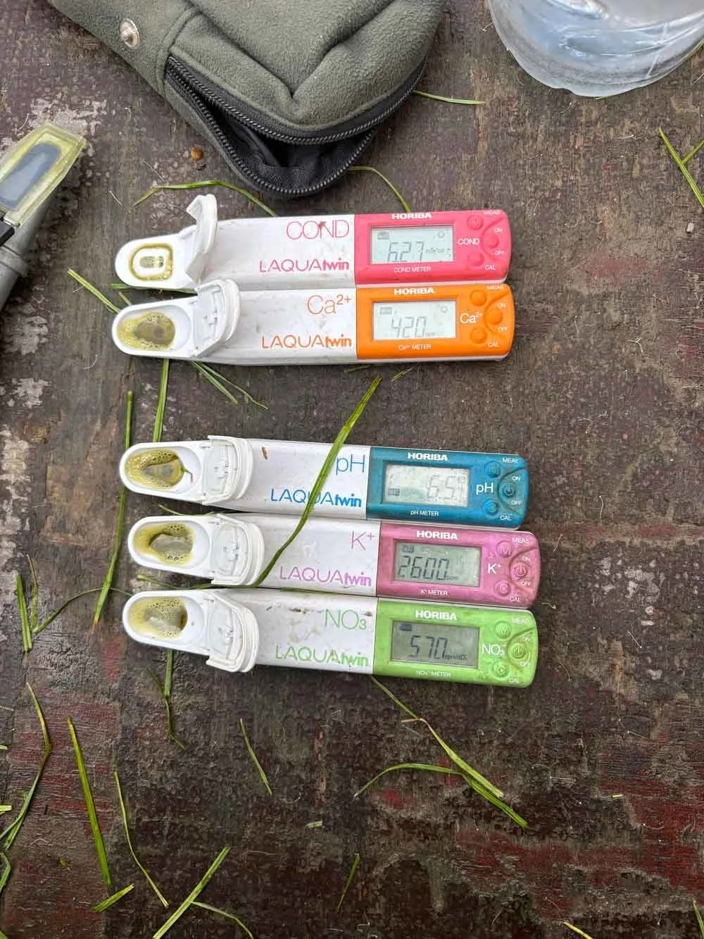 Water testing meters displaying pH 6.53, nitrate 57.0, and other chemical levels on a wooden surface with grass blades around.