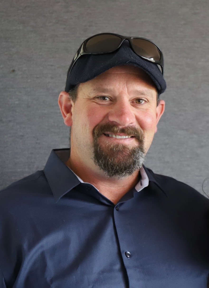 Miah Smith Agrownomics Soil Consultant, smiling wearing a navy blue shirt, a black cap with sunglasses on top, standing against a gray background.