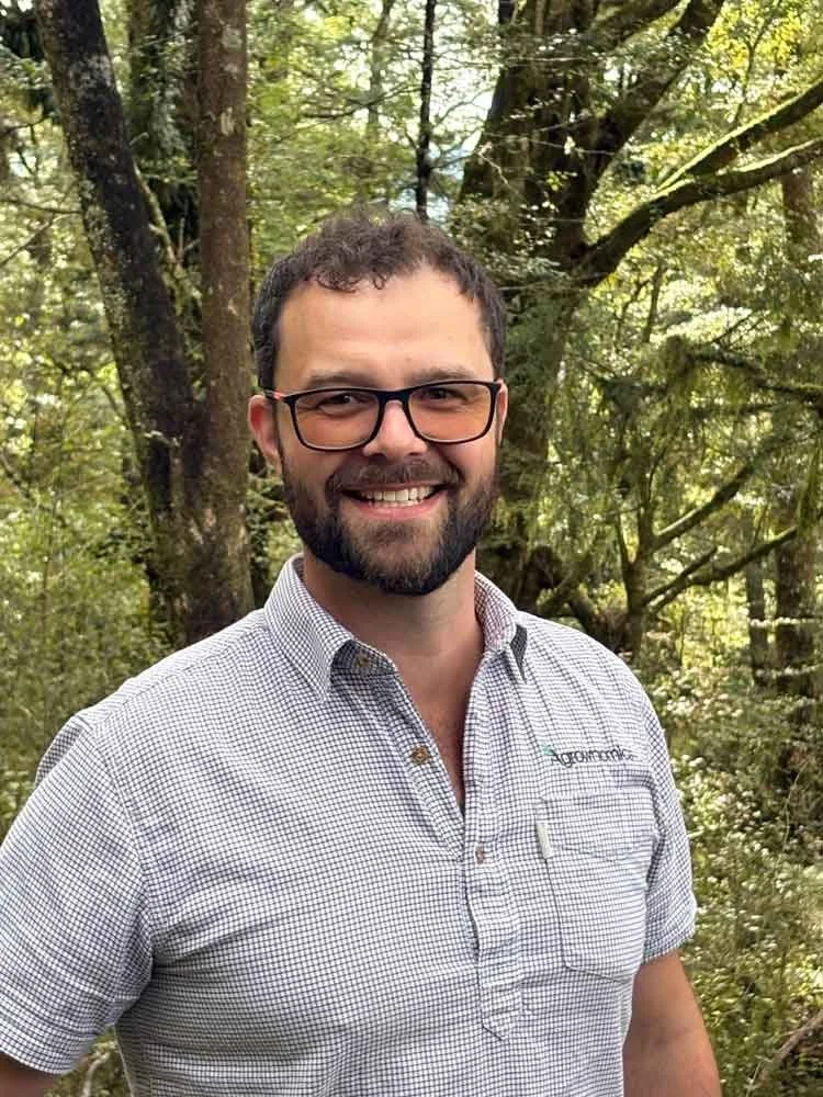 Canaan Ahu Agrownomics CEO & Founder, smiling outdoors in a wooded area with green trees in the background.