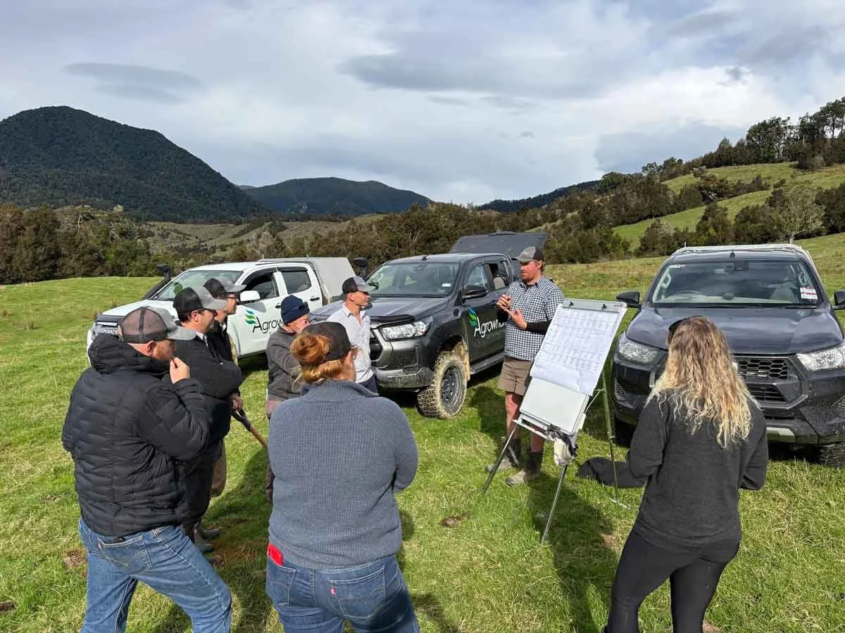 Group of farmers standing outdoors on a grassy field listening to one of the Agrownomics team give a presentation, with multiple vehicles and hills in the background.