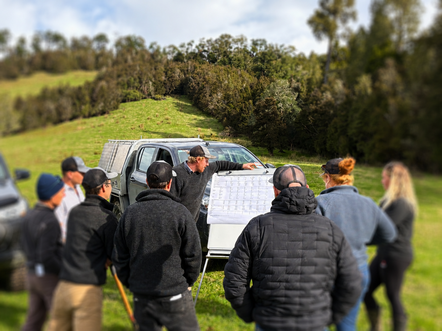 A group of people gathered outdoors on a farm, listening to an Agrownomics team member giving a presentation, with a hillside and trees in the background.