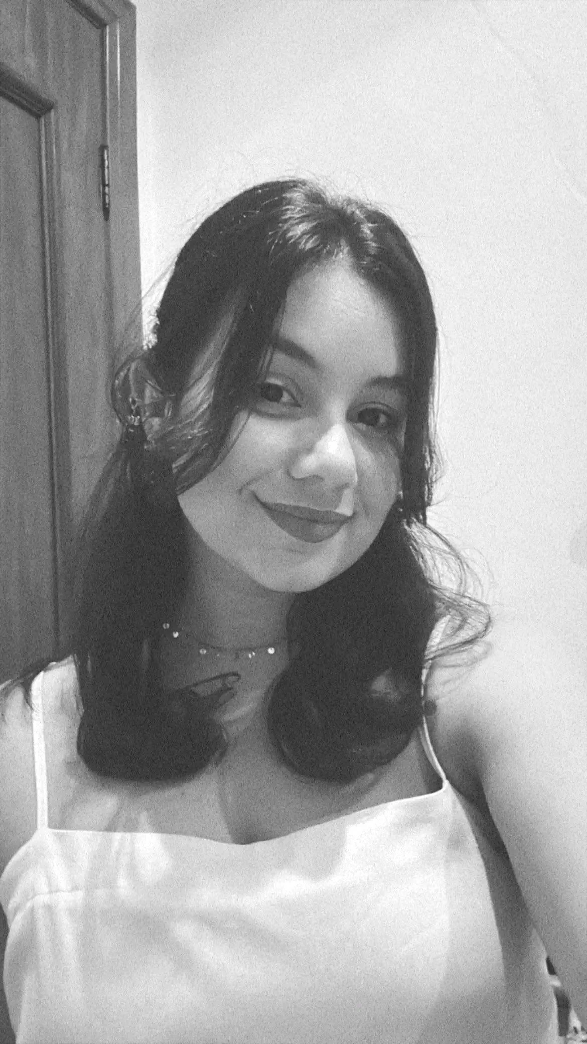 Retrato em preto e branco de uma mulher com cabelo escuro, tamanho médio e batom escuro, sorrindo levemente, usando um vestido branco fino e um colar delicado, com uma porta de madeira ao fundo.