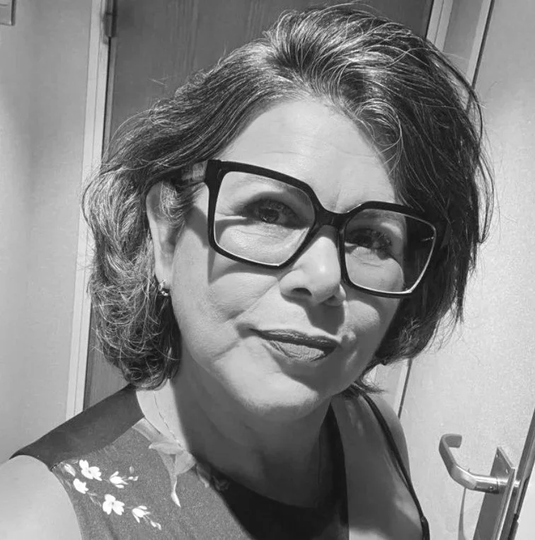 Mulher com cabelo curto, usando óculos grandes de armação escura, sorriso sutil e roupa de estampa floral, posando para foto em ambiente interno