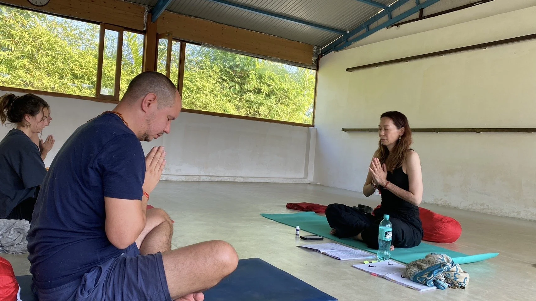 A group of people sitting on yoga mats in a meditation class, with hands in prayer position, indoors with large windows showing green trees outside.