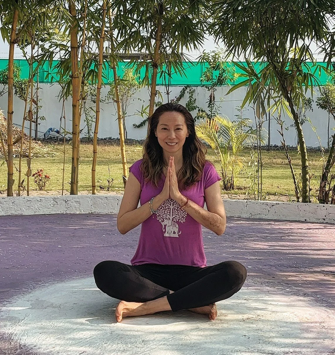 A woman practicing yoga outdoors on a purple mat, sitting cross-legged and holding her hands in prayer position, with a background of bamboo and plants.