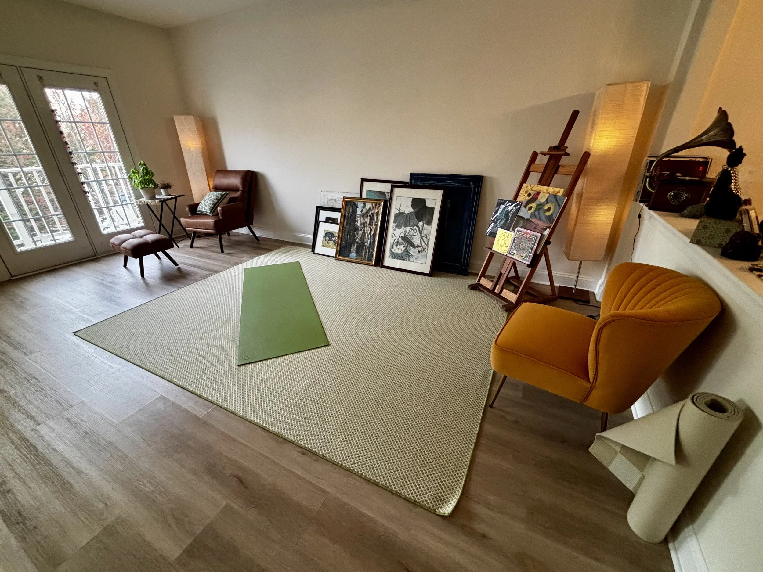 Living room with a green yoga mat on a large beige rug, yellow armchair, pink armchair with ottoman, side table near glass doors with plants, and artwork leaned against the wall.
