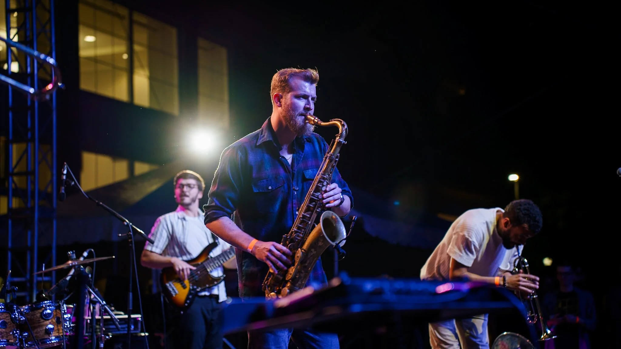 Band performing live on stage with a saxophonist in the center, a guitarist to the left, and a trumpet player to the right, in an outdoor night setting.